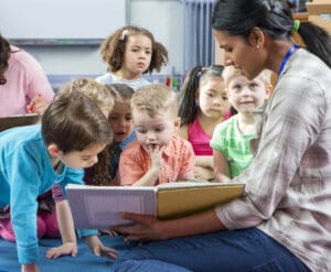 emale teacher giving a lesson to nursery students. They are sitting on the floor and there is a teacher taking notes.